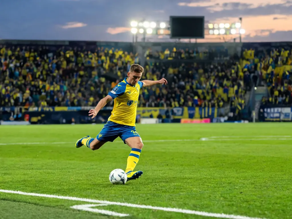Piłkarz Hellas Verona w żółto-niebieskim stroju drybluje piłkę na oświetlonym stadionie, w tle kibice w klubowych barwach.