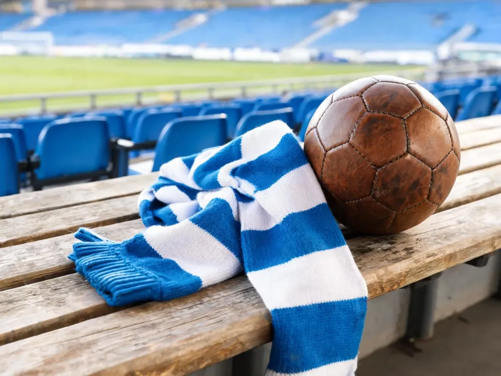 Niebiesko-biały szalik Lecha i stara piłka na ławce stadionu, w tle puste trybuny i murawa przed historycznymi meczami